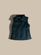 An Andrew Marc black diamond-quilted mixed-media vest with a stand-up collar and front zipper, shown flat against a solid beige background with a soft shadow.
