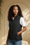A woman wearing an Andrew Marc black diamond-quilted mixed-media vest over a white short-sleeve shirt and blue jeans, standing in front of a beige arched wall with soft shadows.