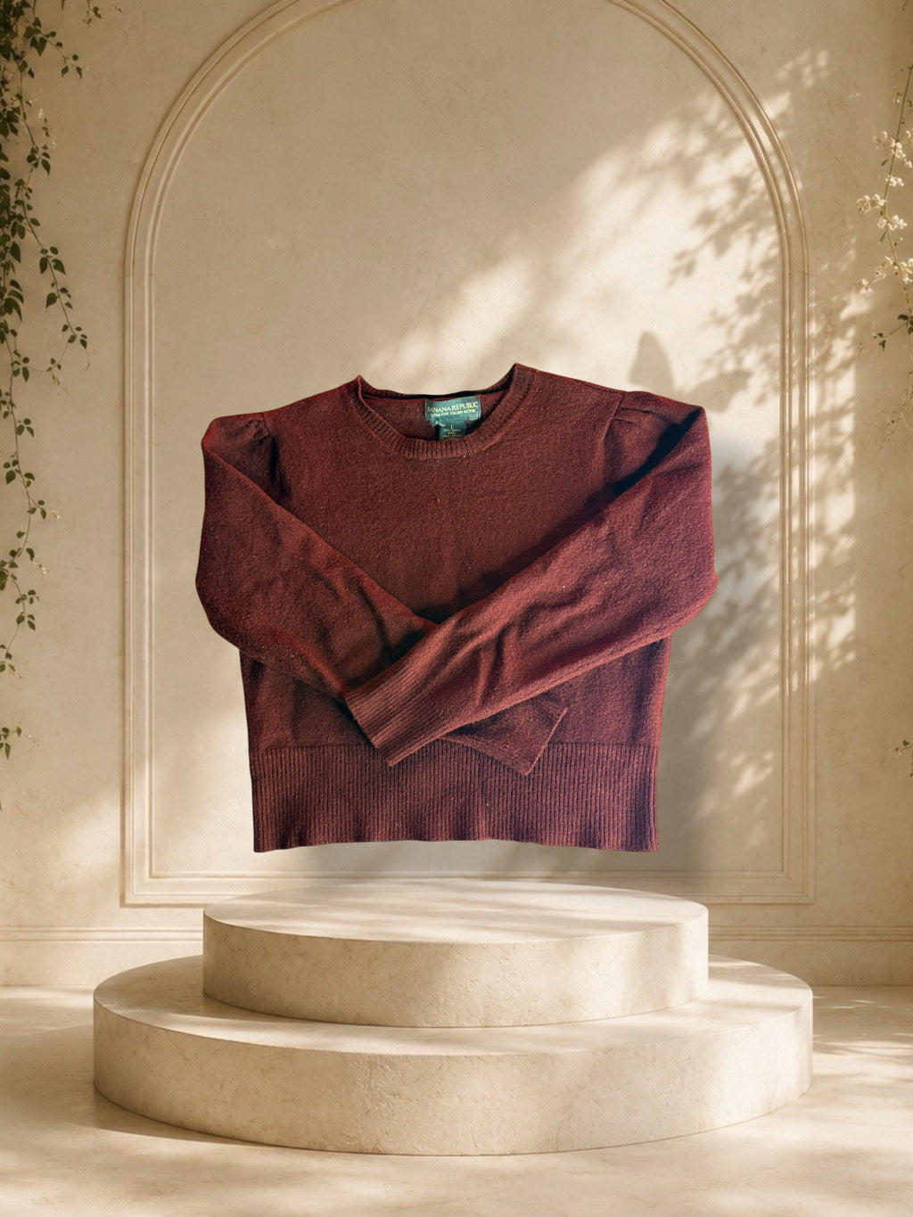 A Banana Republic Italian Merino puff-shoulder sweater detail is displayed on a cream-colored two-tiered pedestal in front of a beige arched wall with soft leaf shadows.