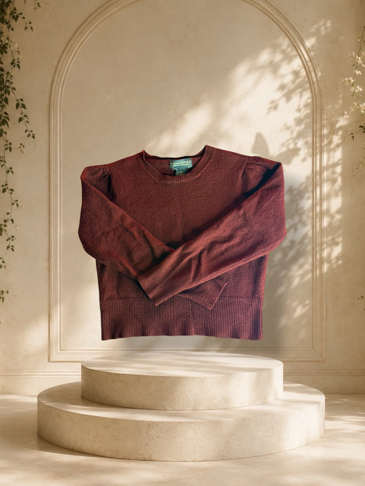 A Banana Republic Italian Merino puff-shoulder sweater detail is displayed on a cream-colored two-tiered pedestal in front of a beige arched wall with soft leaf shadows.