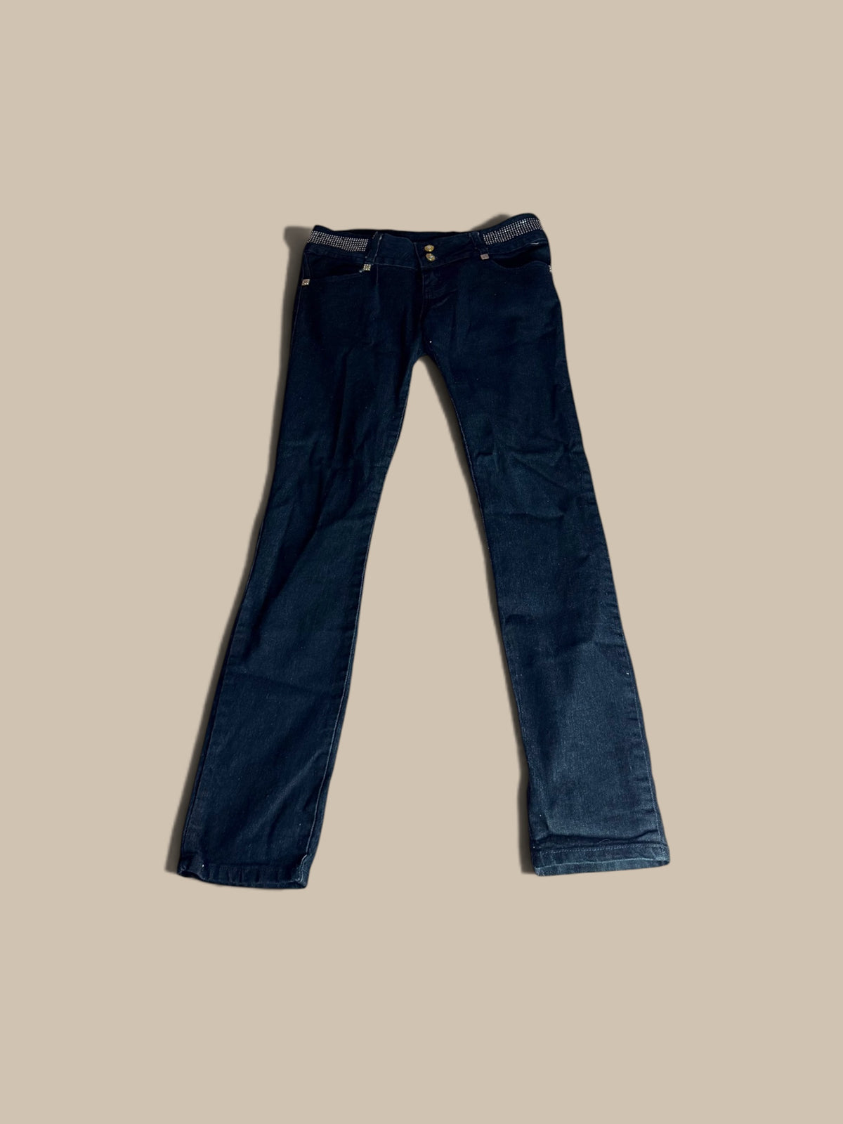 A pair of dark indigo B.B. Jeans rhinestone waistband skinny jeans laid flat against a solid beige background.