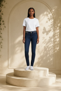 A model stands on two round pedestals in a warm, sunlit studio setting. She is wearing a plain white short-sleeve crewneck t-shirt, B.B. Jeans rhinestone waistband skinny jeans, and white sneakers.