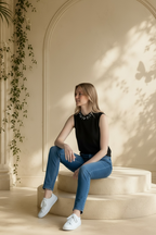 A woman sits on cream-colored stone steps in a sunlit architectural setting, wearing a sleeveless Carmen Marc Valvo black embellished knit shell top with a pearl-like collar detail, blue jeans, and white sneakers.