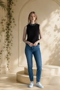 A woman stands in a warm beige interior with arched wall details and soft shadows, wearing a sleeveless Carmen Marc Valvo black embellished knit shell top with a jeweled collar, blue jeans, and white sneakers.