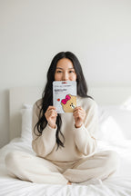 A woman with long black hair sits cross-legged on a white bed, holding a CHOCO printed sheet mask from The Crème Shop x LINE FRIENDS in front of her face. She is wearing a cream-colored knit pajama set.