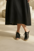 A person wearing a long black skirt and Free People Gable black gladiator espadrille slide sandals with braided jute soles, standing on a light beige floor in a sunlit interior.