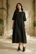 A woman stands in a sunlit architectural interior wearing a loose black midi dress with short sleeves and Free People Gable black gladiator espadrille slide sandals.