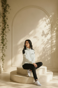 A woman sits on a round beige pedestal in a sunlit room with an arched wall and leaf shadows. She is wearing an I.N.C. International Concepts faux-leather trim full-zip knit jacket, black pants, and white sneakers.