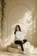 A woman sits on a round beige pedestal in a sunlit room with an arched wall and leaf shadows. She is wearing an I.N.C. International Concepts faux-leather trim full-zip knit jacket, black pants, and white sneakers.