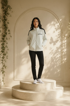 A woman stands on a round stone platform in a warm, sunlit architectural setting, wearing an  I.N.C. International Concepts faux-leather trim full-zip knit jacket with dark shoulder panels, black pants, and white sneakers.