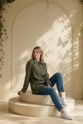 Model sits on pedestal wearing Joan Vass USA vintage herringbone knit shirt jacket in olive green and charcoal, blue jeans, and white sneakers.