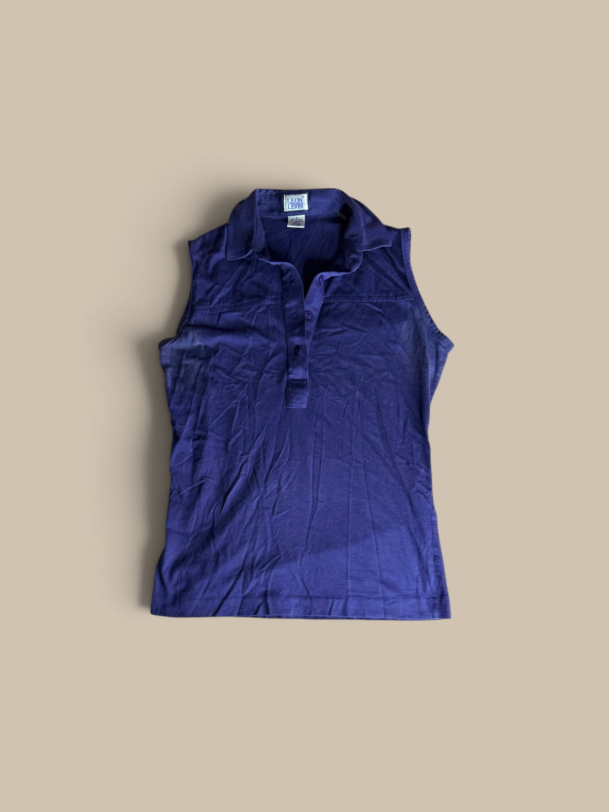 A sleeveless, Leon Levin deep purple indigo polo shirt, laid flat against a solid beige background.