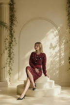 A woman sits on cream-colored steps in a sunlit architectural setting, wearing a Nessa Samantha crimson red and black houndstooth knit sheath dress with decorative buttons and black pointed heels.