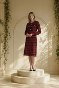 A woman stands on a round cream pedestal in a sunlit interior with arched wall molding and hanging greenery. She is wearing a Nessa Samantha crimson red and black houndstooth knit sheath dress modeled standing and black pointed heels.