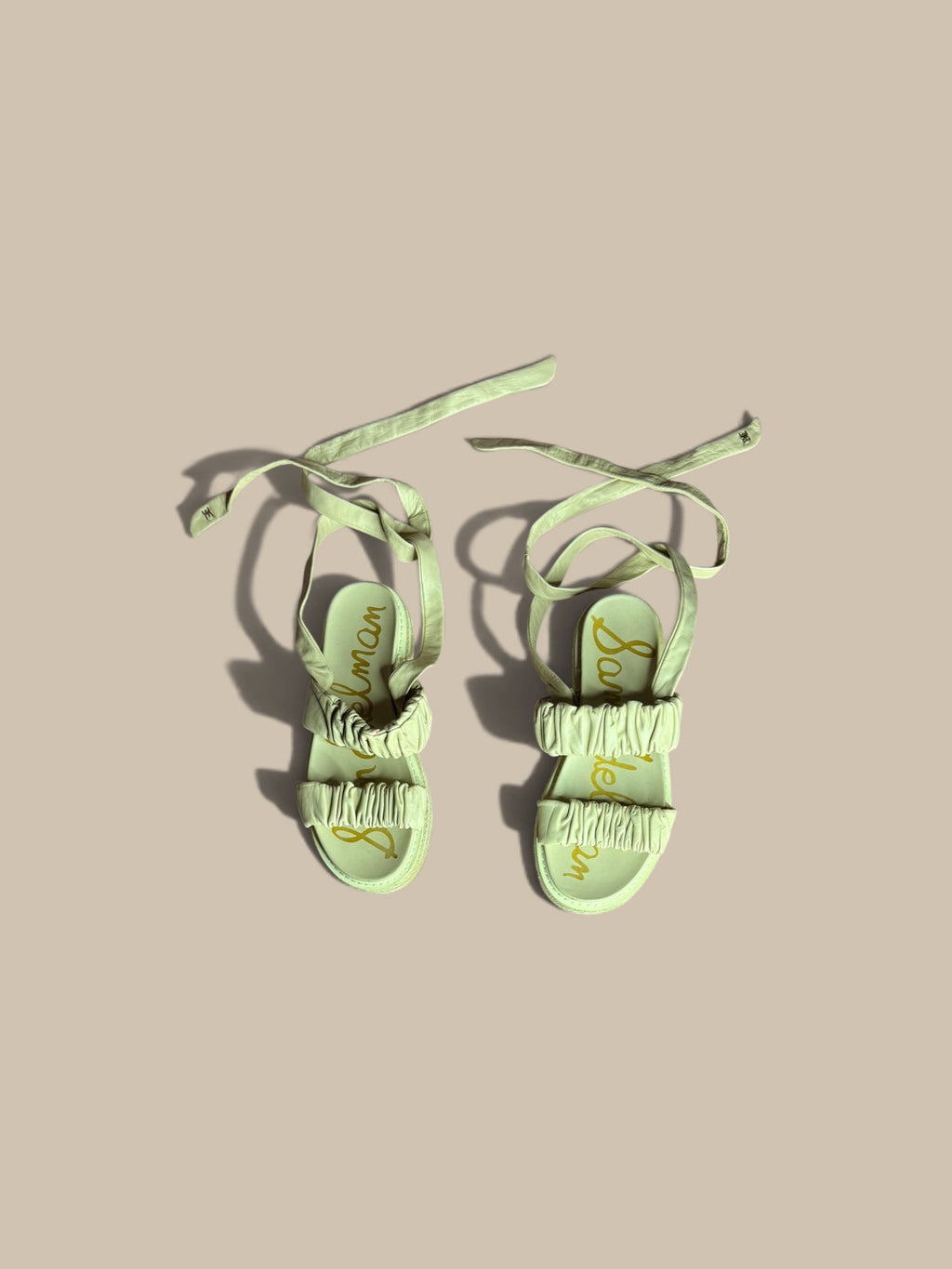 A pair of Sam Edelman Velma ruched ankle-tie espadrille sandals with ruffled straps and long tie-backs, featuring gold script lettering on the insoles, displayed on a solid beige background.
