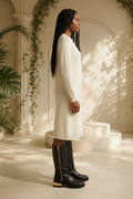 A woman stands in profile wearing a long cream knit dress and Vince Camuto Winxie black leather fold-over moto boots with gold-toned zipper details, posed in a sunlit architectural setting with columns, plants, and a small raised platform.