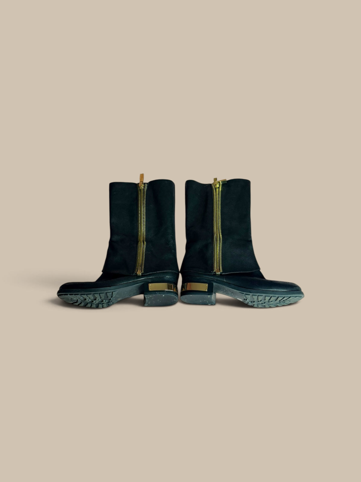 A pair of Vince Camuto Winxie black leather moto boots showing outer zipper sides against a solid beige background.